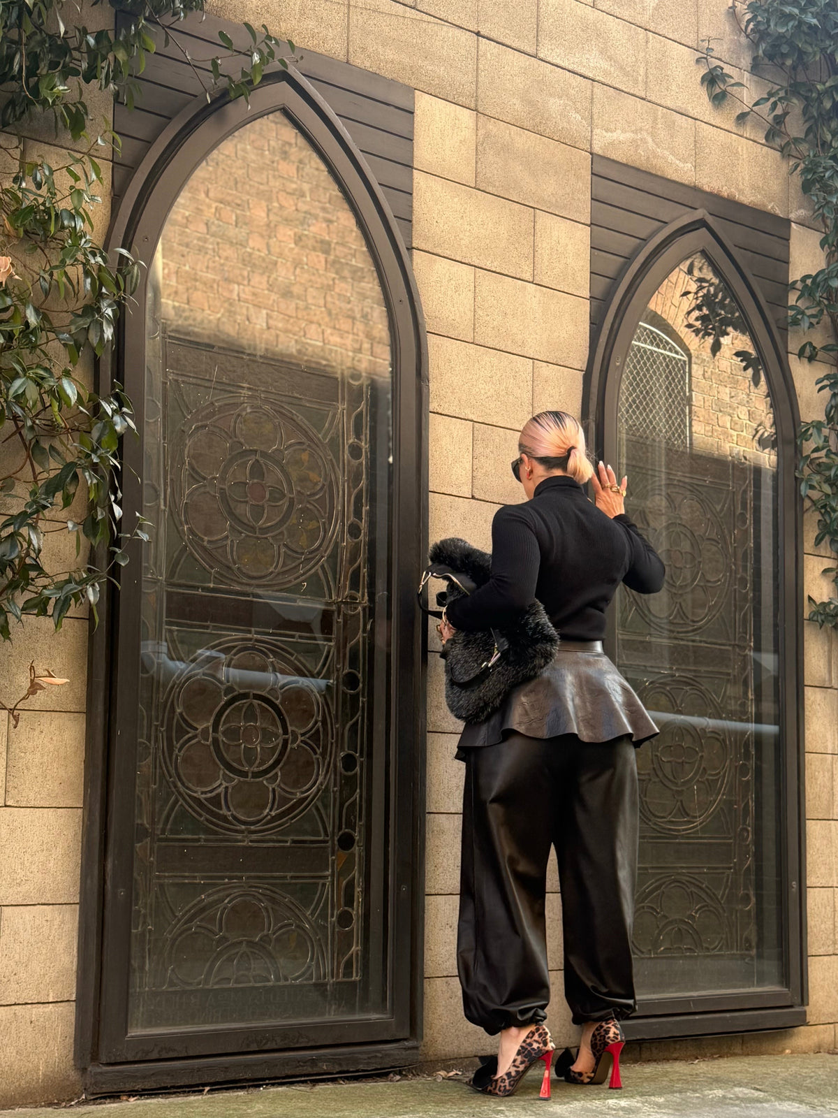 A person stands with their back to the camera in front of two large arched gothic-style glass doors set in a beige stone wall. The person is wearing a black long-sleeve top, a dark metallic peplum-style waistcoat, loose black trousers, and red high-heeled shoes, while holding a black furry handbag.